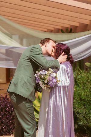A couple shares a romantic kiss outdoors with the bride wearing lavender and holding a purple floral bouquet.