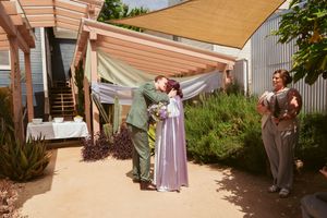 People in formal attire gather under a wooden pergola with beige shade sails on a sunny afternoon.