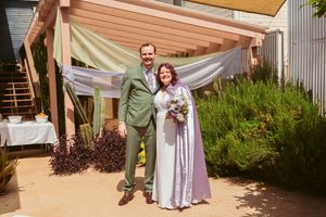 The newlyweds pose for a wedding photo under a tan canopy decorated with sheer fabric and garden landscaping.
