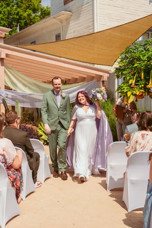 A wedding couple walking down the aisle surrounded by guests in white chairs under a canopy with desert decor.