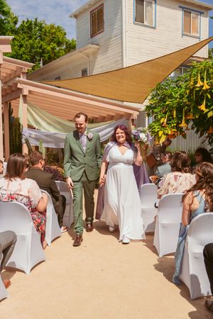 An outdoor backyard wedding ceremony under a beige shade sail with guests seated in white chairs.