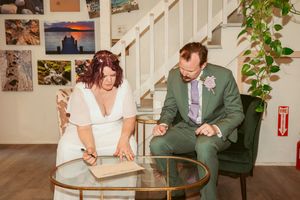 Two people sit at a glass coffee table indoors with photo collages displayed on the wall behind them.