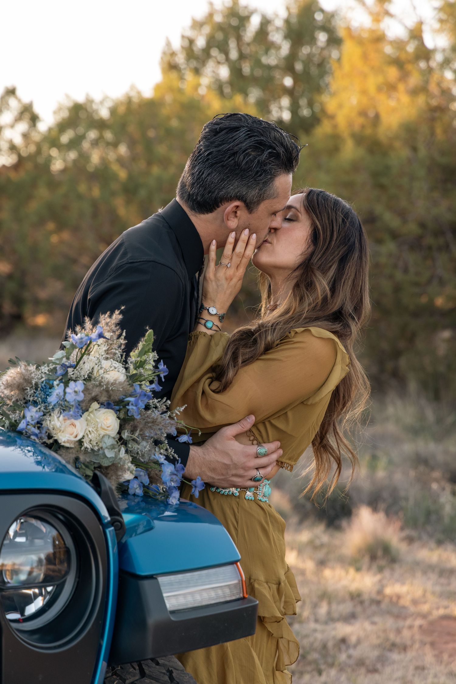 A romantic couple share a kiss beside a blue vintage car as they hold a delicate bouquet of blue and white flowers at sunset.