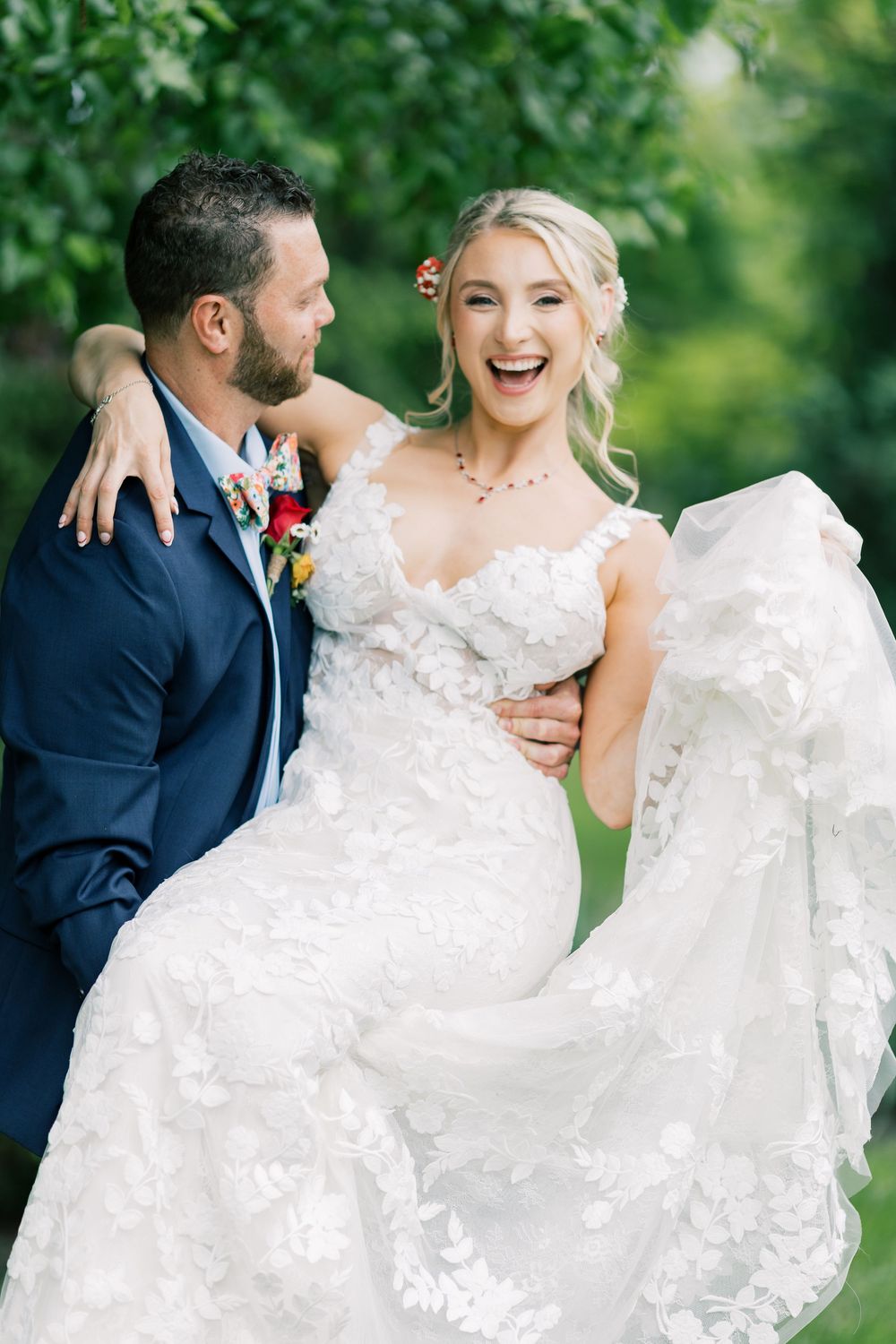 A playful wedding photo showing a groom lifting his bride in a lace dress amidst greenery in Orange County, New York.