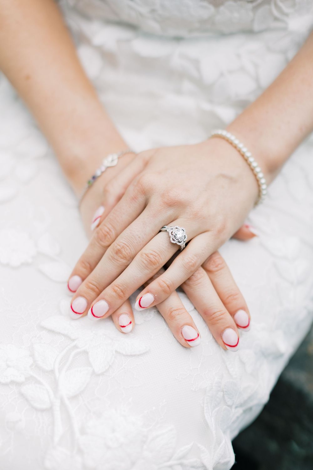 Close-up of bride's hands showing French manicure, wedding ring and pearl bracelet on white lace dress in Orange County, New York.