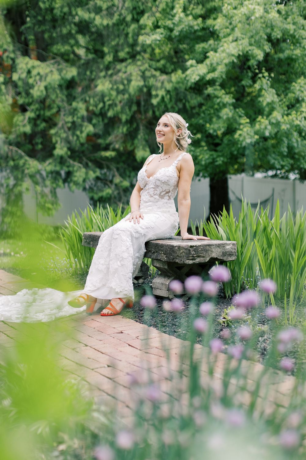 Bride in lace gown sitting serenely by a garden pond surrounded by purple flowers and greenery in Orange County, New York.