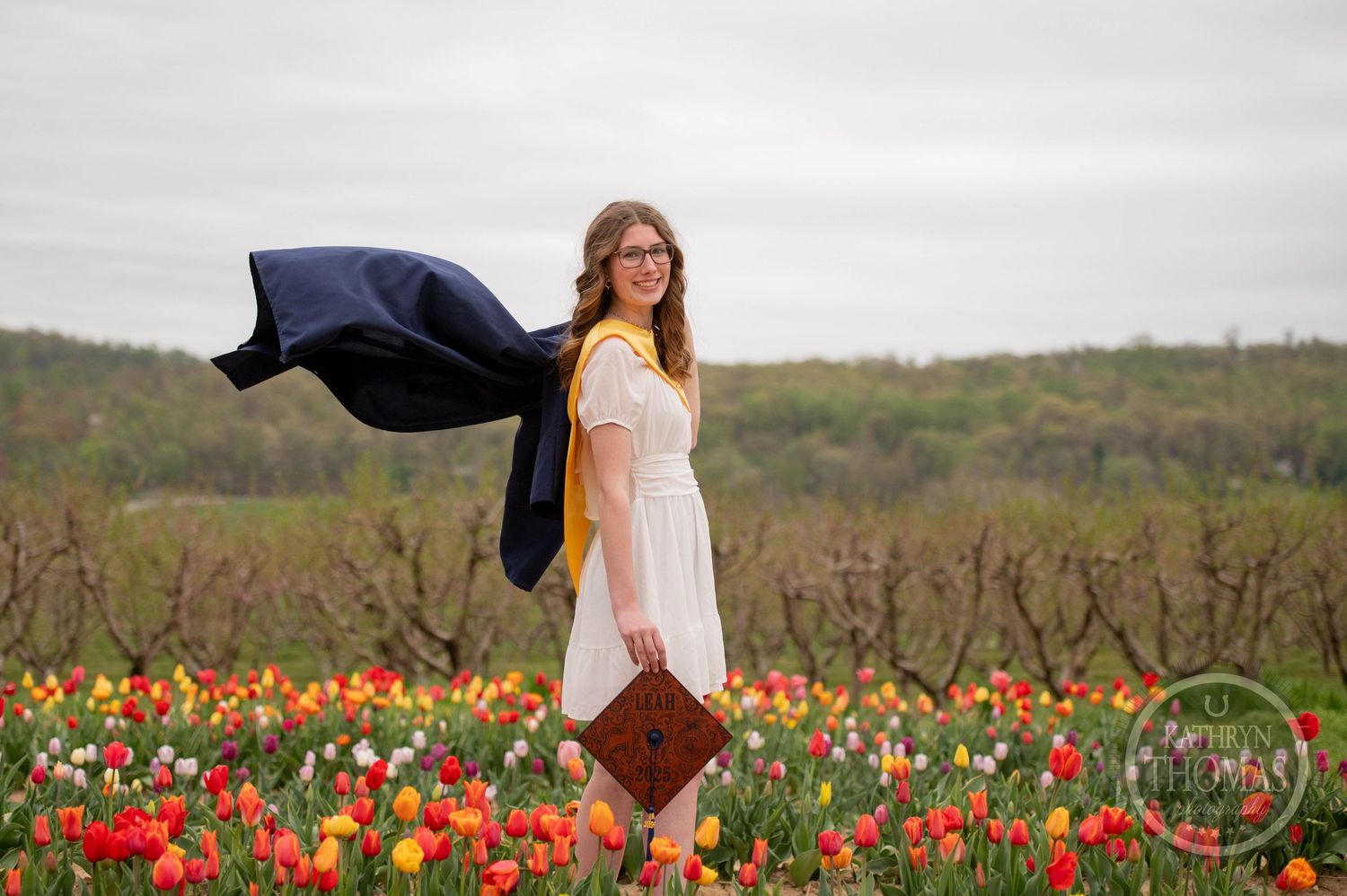 Person in white dress stands in blooming tulip field with flowing navy jacket against cloudy sky.