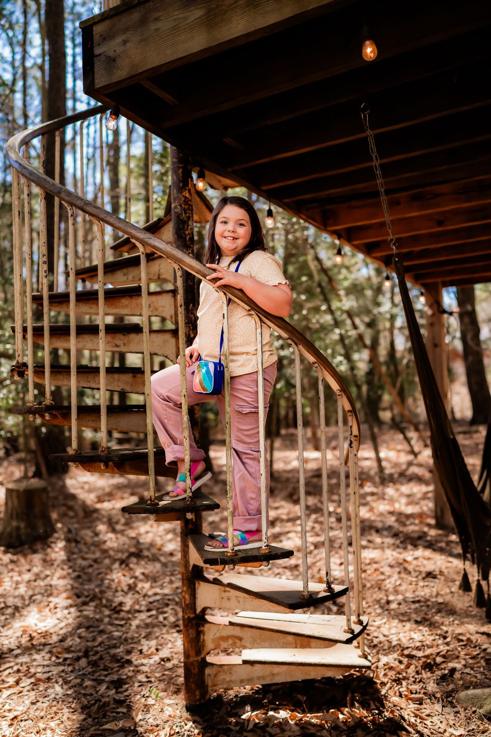 Someone stands on a rustic spiral wooden staircase in the woods wearing pink pants and a white top.