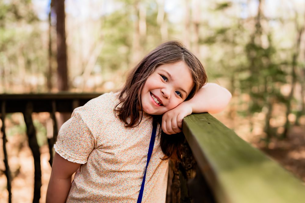 Someone in a beige shirt leans against a wooden railing in a forest setting with a cheerful expression.