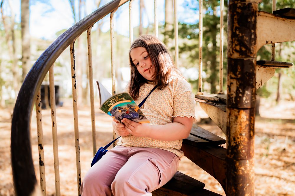 Person in a beige sweater sitting outdoors on a curved swing while reading a book in a wooded setting.