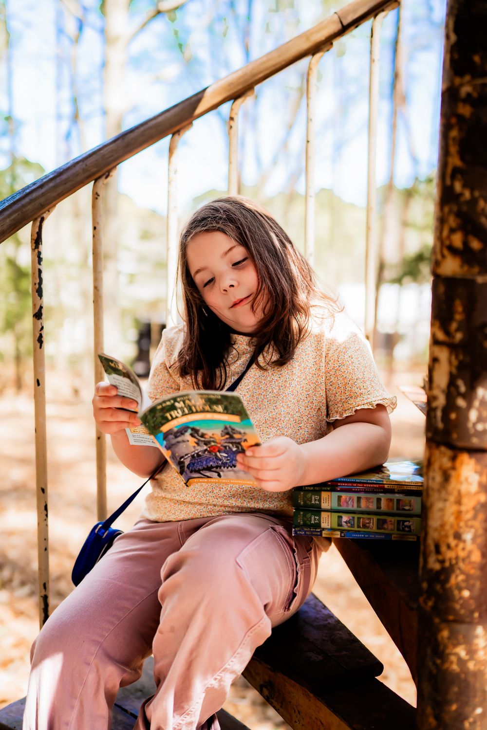 Person reading outdoors on wooden steps with stack of books beside them on a sunny autumn day.