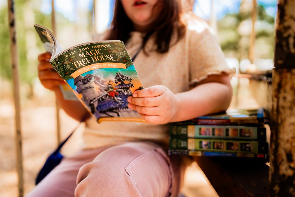Someone reads a Harry Potter book in pink pants while sitting outdoors with a stack of other books nearby.