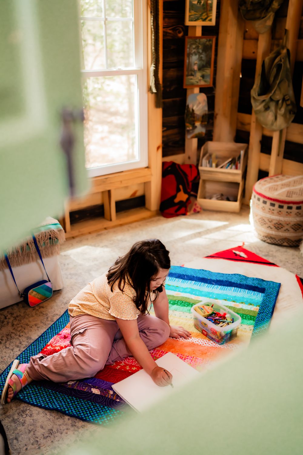 Young person in peach-colored clothing sits on a colorful striped rug in a sunlit room with craft supplies.