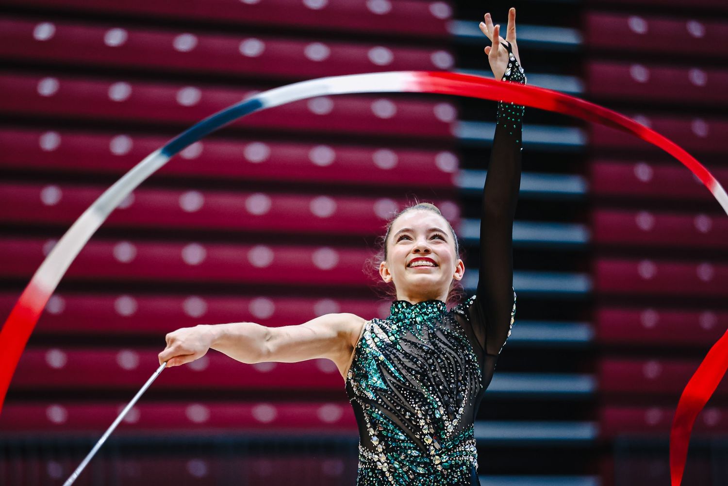 A gymnast in a blue and black leotard performs with arms raised in a bright indoor gymnasium.
