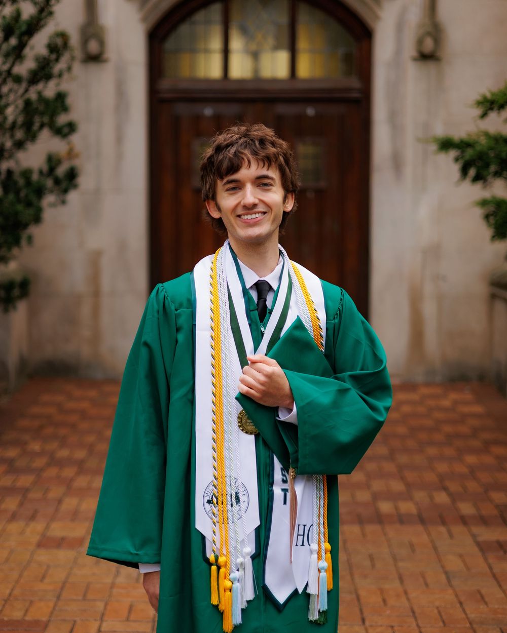 Smiling graduate in green cap and gown with honor cords poses outdoors at graduation ceremony.