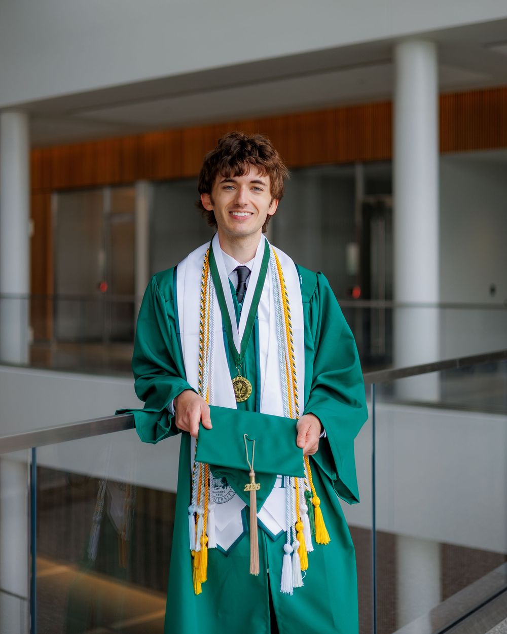 Smiling graduate in green cap and gown with gold honor cords poses indoors at graduation ceremony.