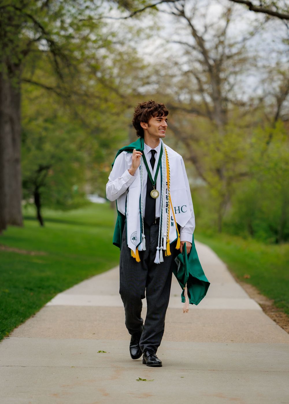 Graduate walks outdoors in green cap and gown regalia, wearing honor cords and medal on a tree-lined path.