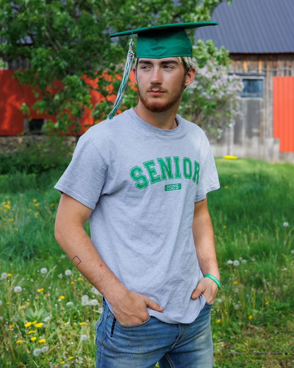 Graduate wearing a green cap and grey Senior t-shirt poses outdoors with rustic barn in background.