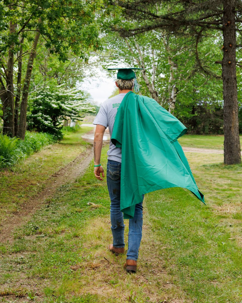 A person wearing a graduation cap and gown walks along a tree-lined path in jeans on a sunny summer day.