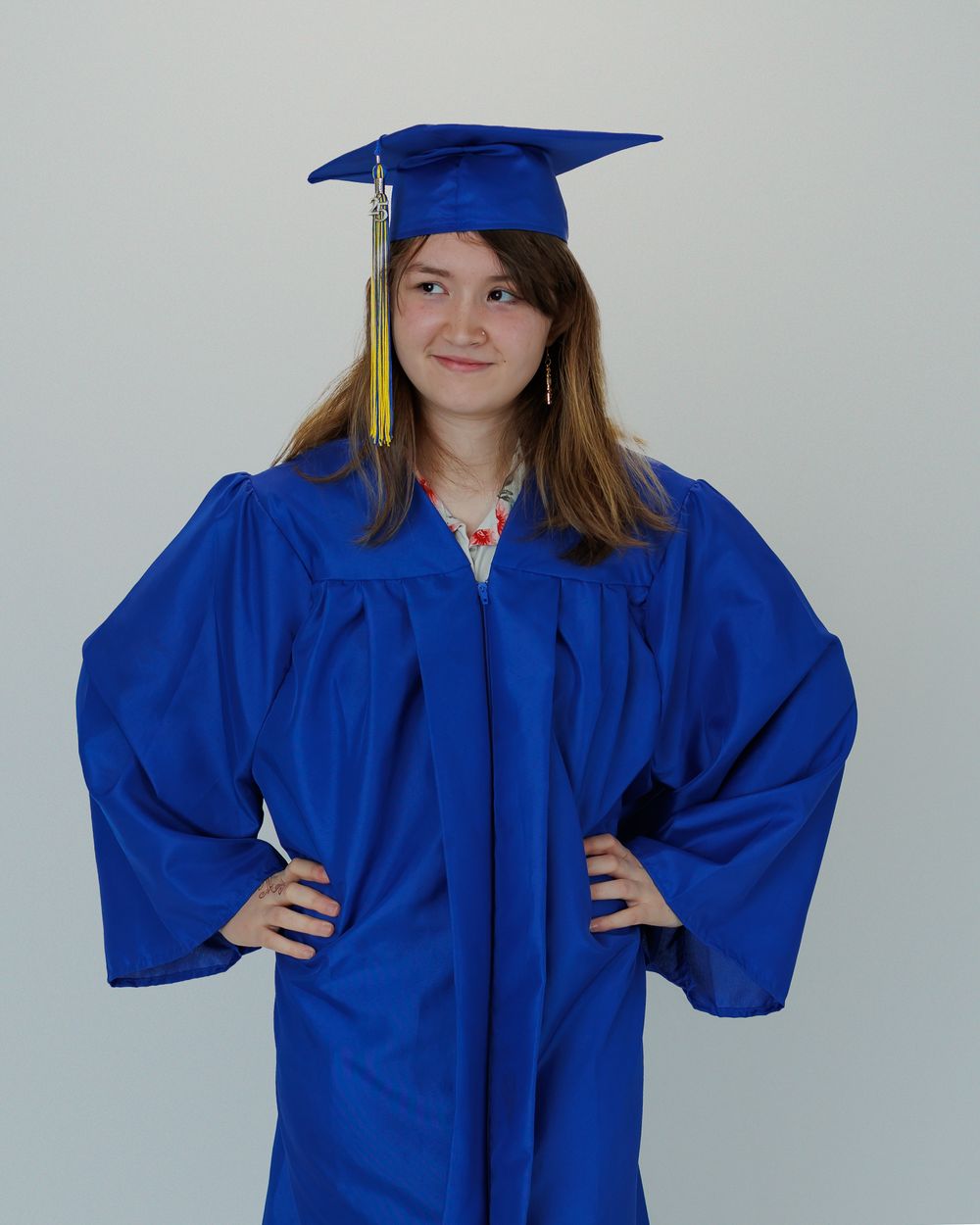 Graduate poses in bright blue cap and gown with gold tassel and hands on hips against neutral background.
