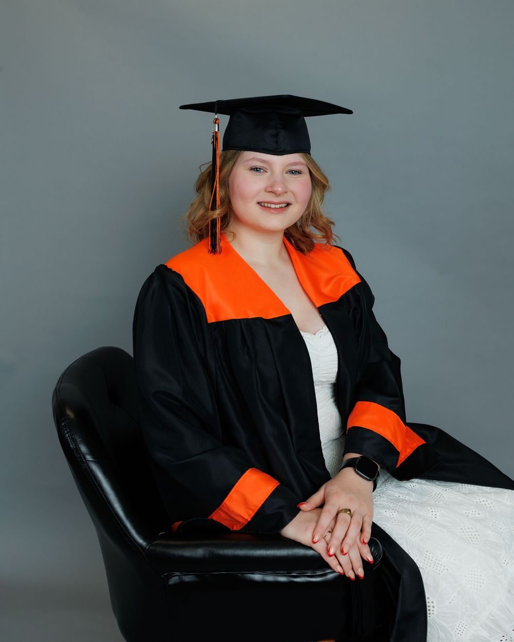 Graduate wearing a traditional black cap and gown with orange stole seated in a leather chair against a gray backdrop.