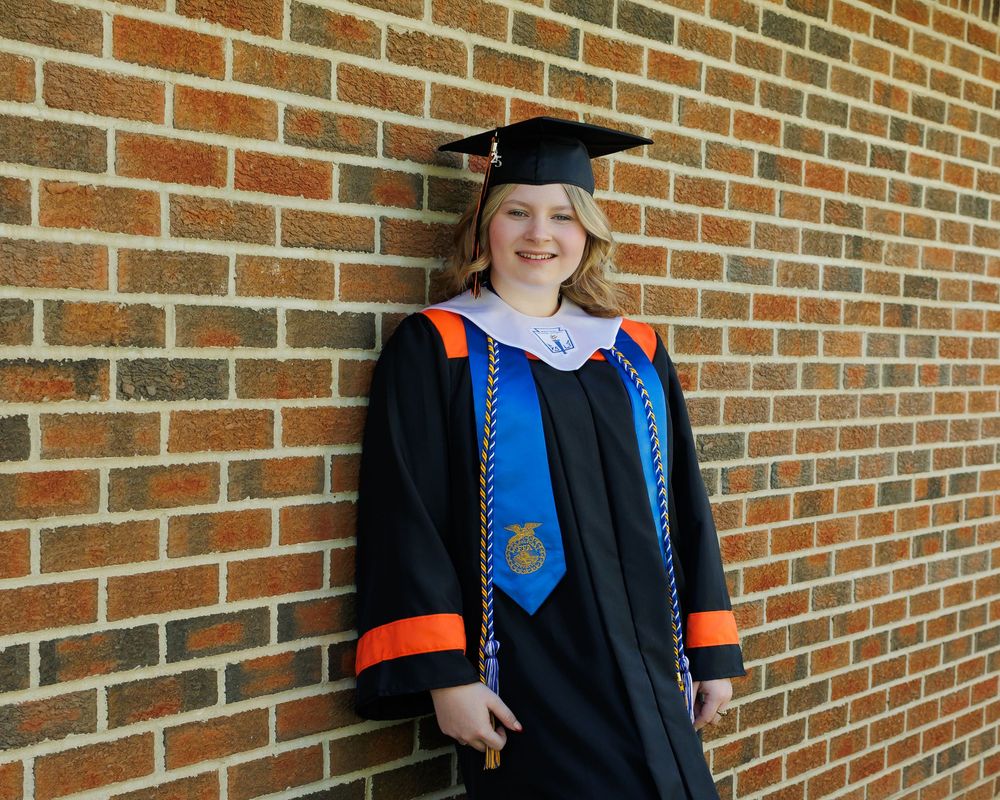 Graduate in black cap and gown with blue stole poses against brick wall for commencement photos.