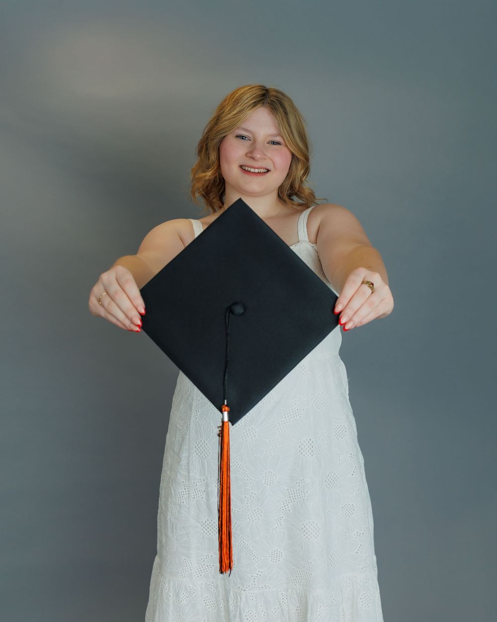 Graduate in white dress holds up mortarboard graduation cap with orange tassel against grey backdrop.