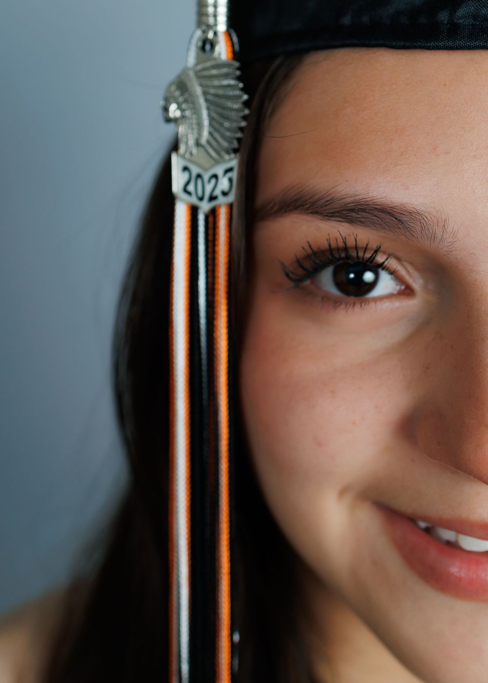 A graduation tassel marked with '2025' hangs next to a closeup partial view showing long eyelashes and a smile.