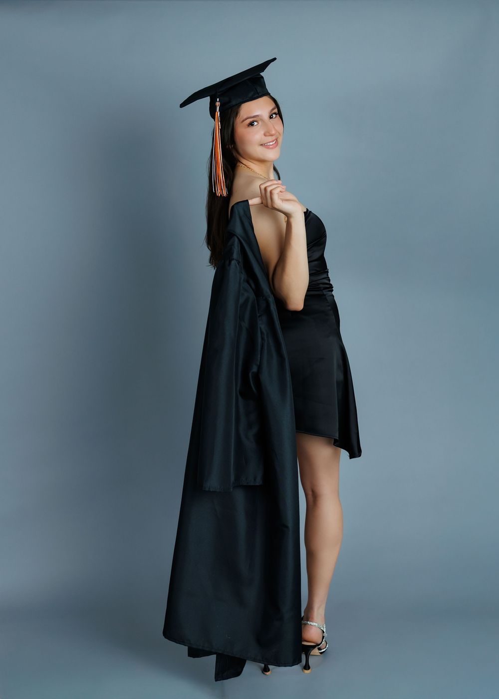 Graduation portrait showing graduate posing in traditional black cap and gown with one leg forward against gray backdrop.
