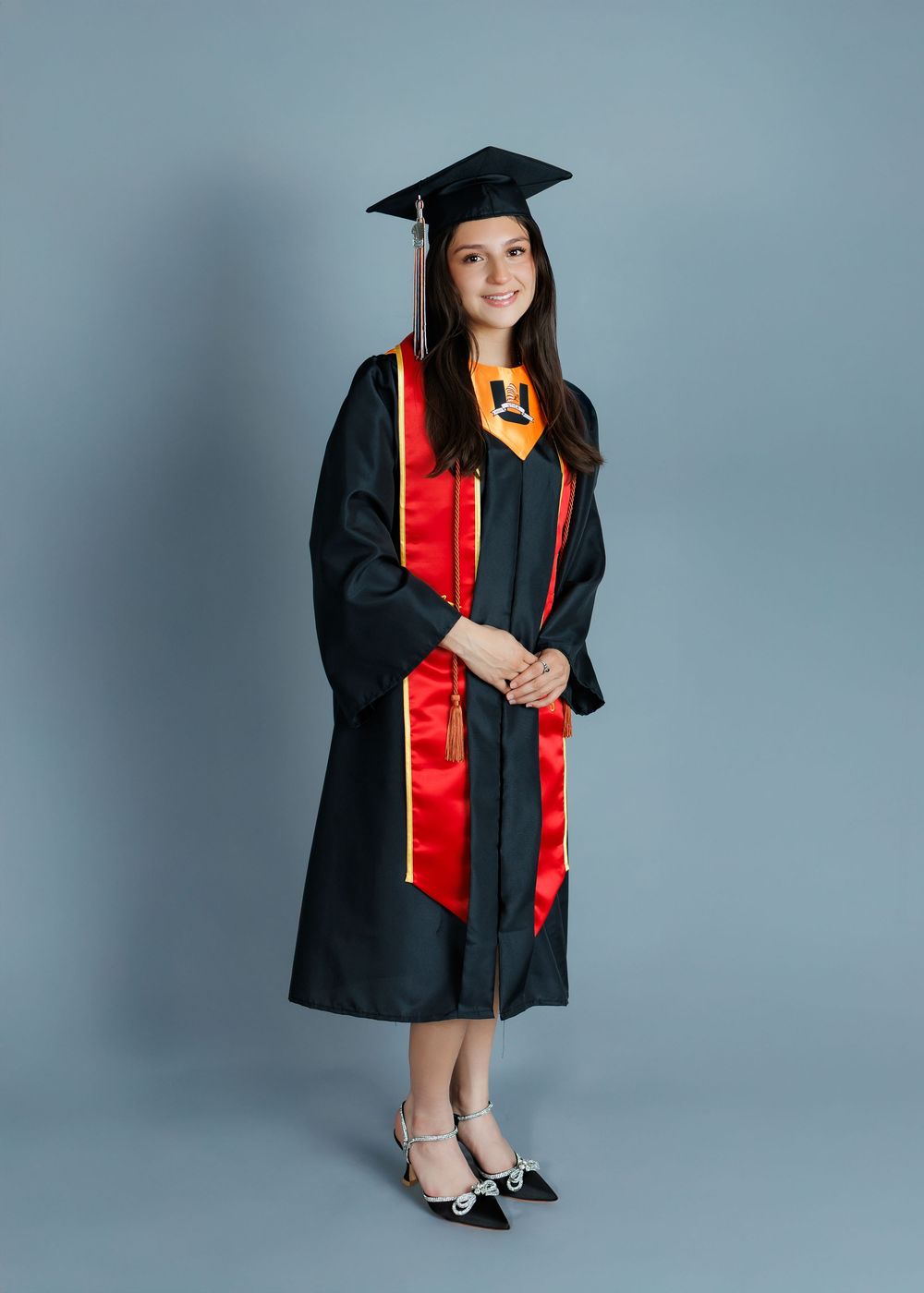 Graduate in black cap and gown with red stole stands against gray background wearing pointed flats.
