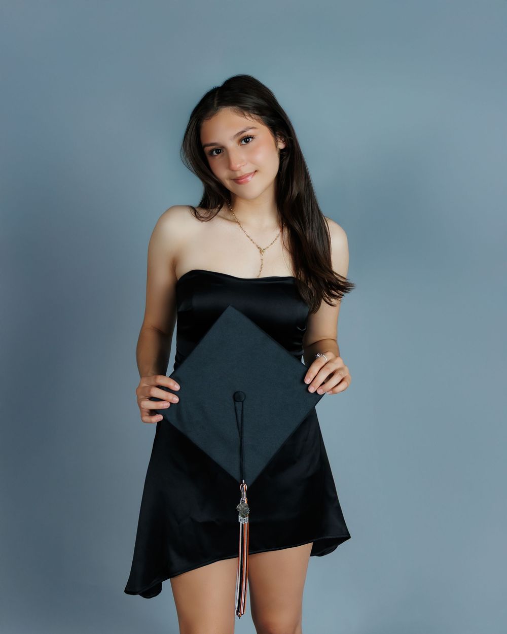 A stylish graduate posing in a black cocktail dress while holding a graduation cap against a blue studio background.