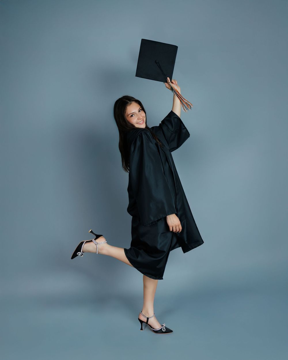 Graduate in traditional black cap and gown jumps joyfully while celebrating graduation against blue backdrop.