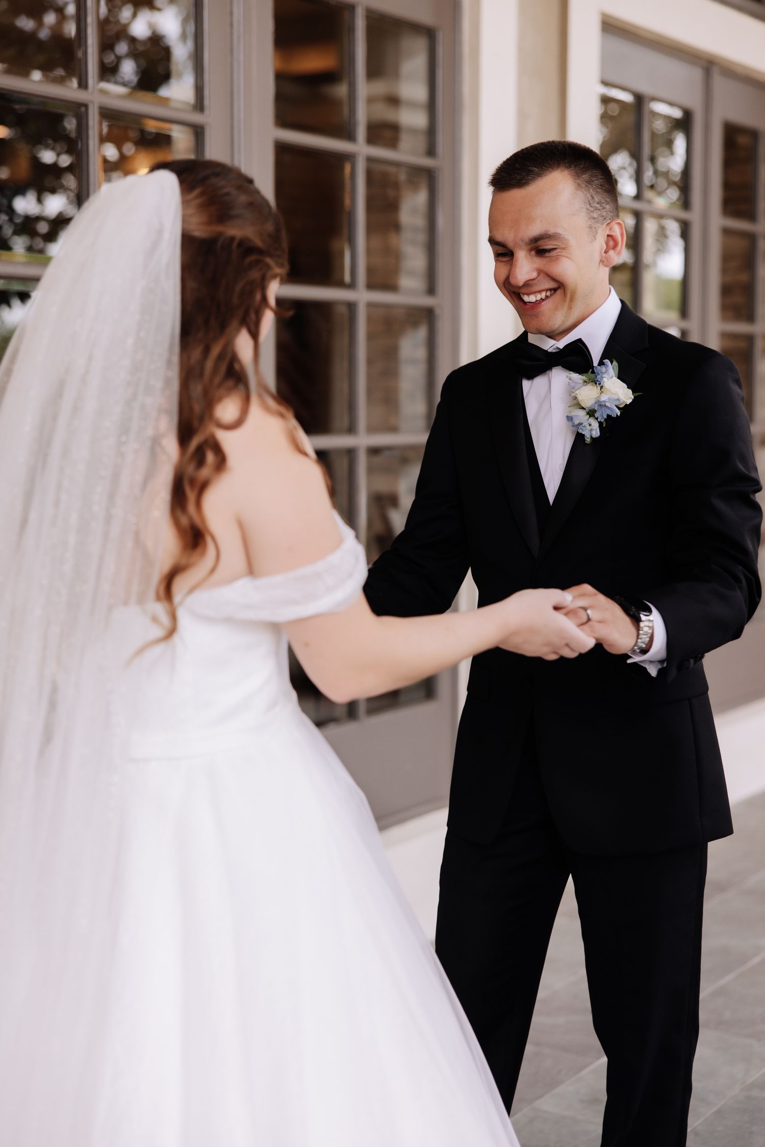 A bride in a white ballgown and veil holds hands with a groom in classic black tuxedo during a joyful first look moment.