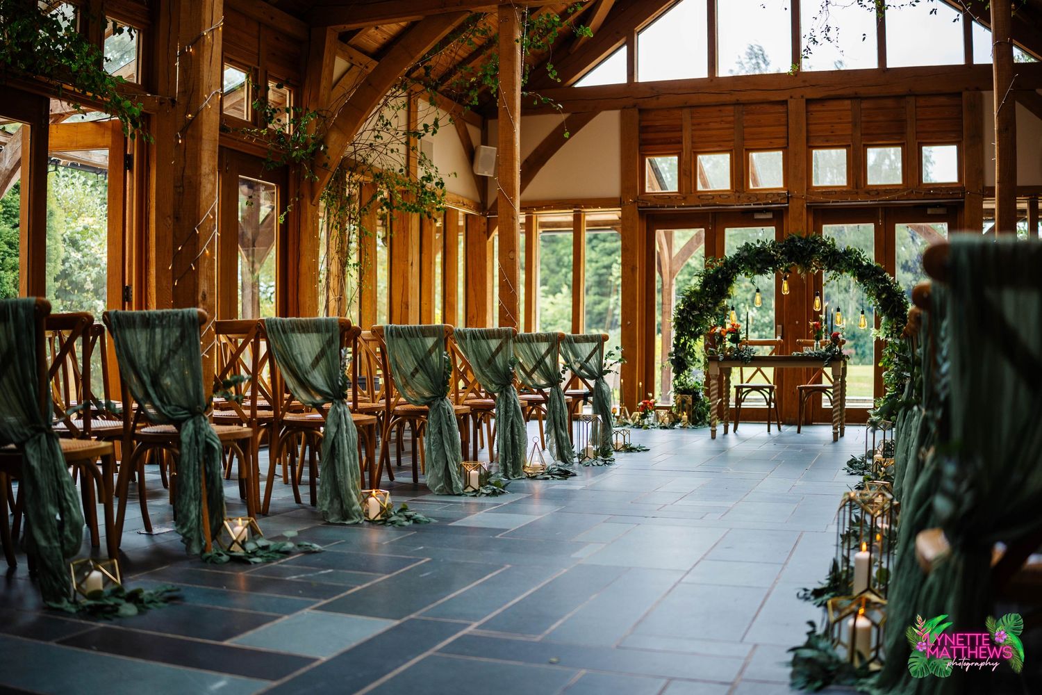 Rustic barn wedding venue with wooden beams, cross-back chairs and floral arch decorations for ceremony setup at The Oaktree of Peover