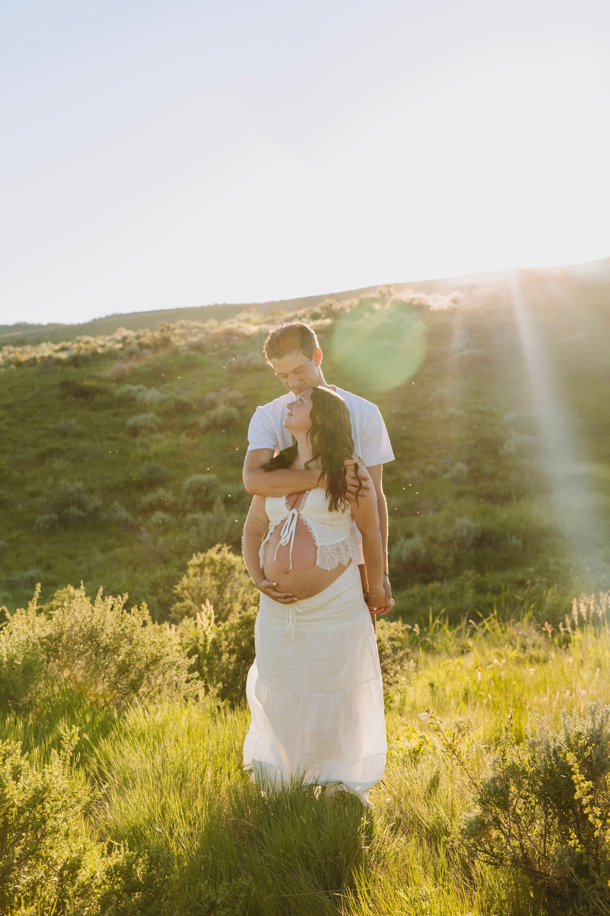 Couple embracing in sunlit green field, pregnant woman in white dress with exposed belly, golden hour lens flare.