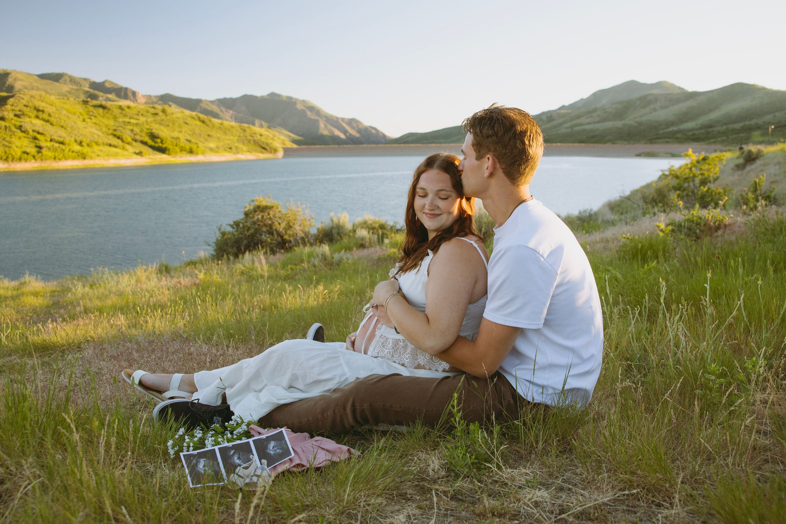 Pregnant couple sitting by a scenic lake in green hills, holding ultrasound photos during a maternity photoshoot outdoors.