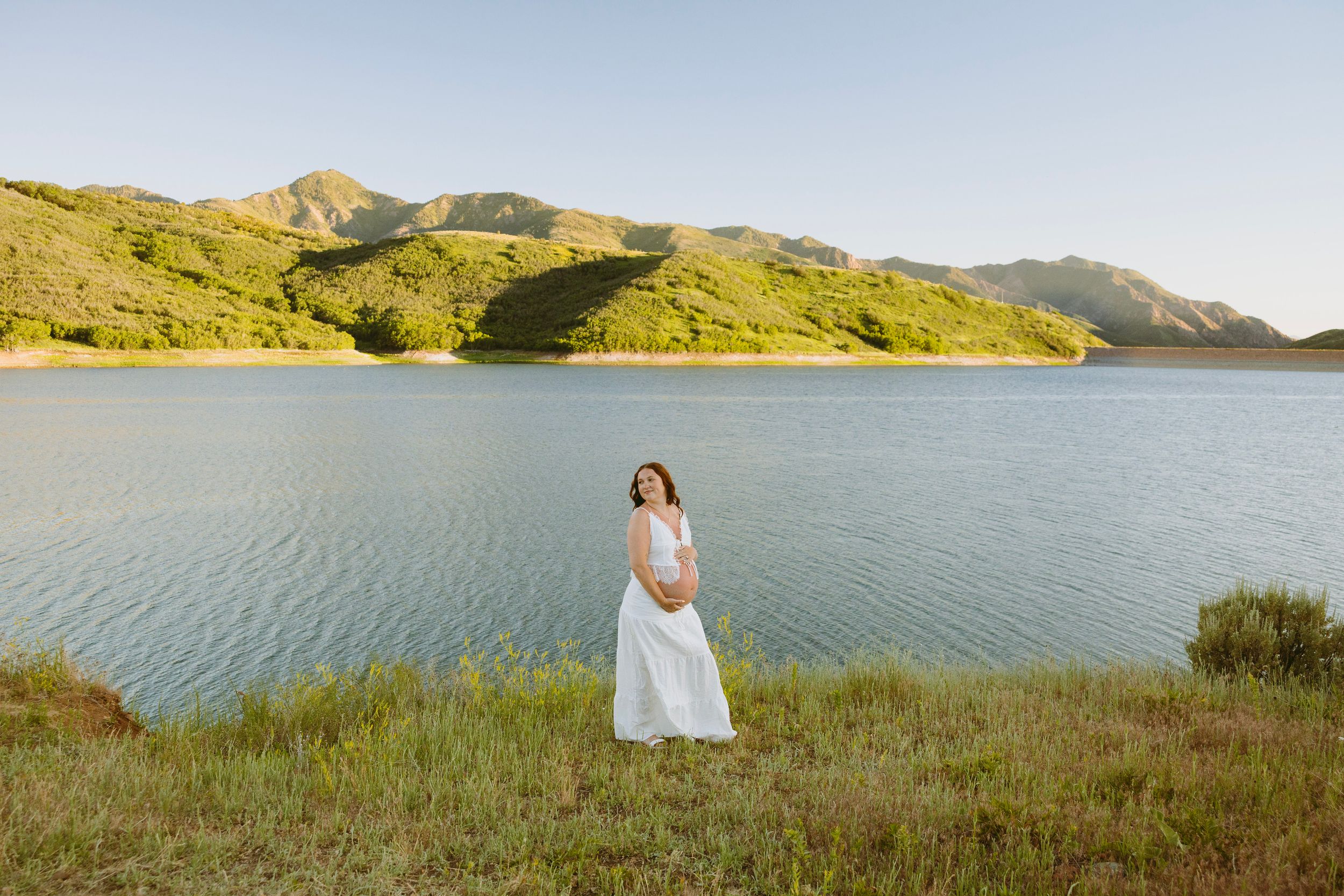 Pregnant woman in white dress standing by a serene lake with lush green hills in the background.