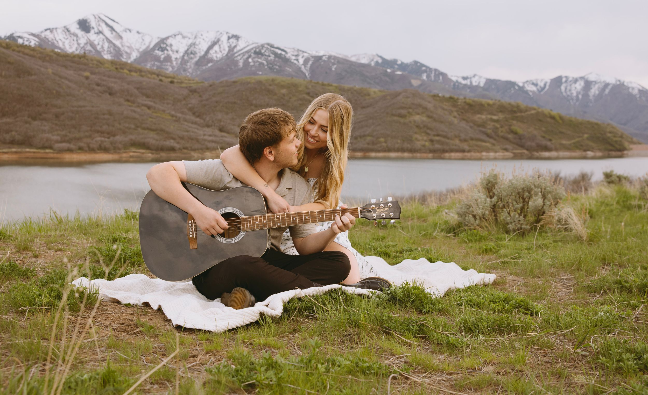 An engaged couple sharing a musical moment with a guitar on a blanket by the lakeside in Salt Lake City, Utah.