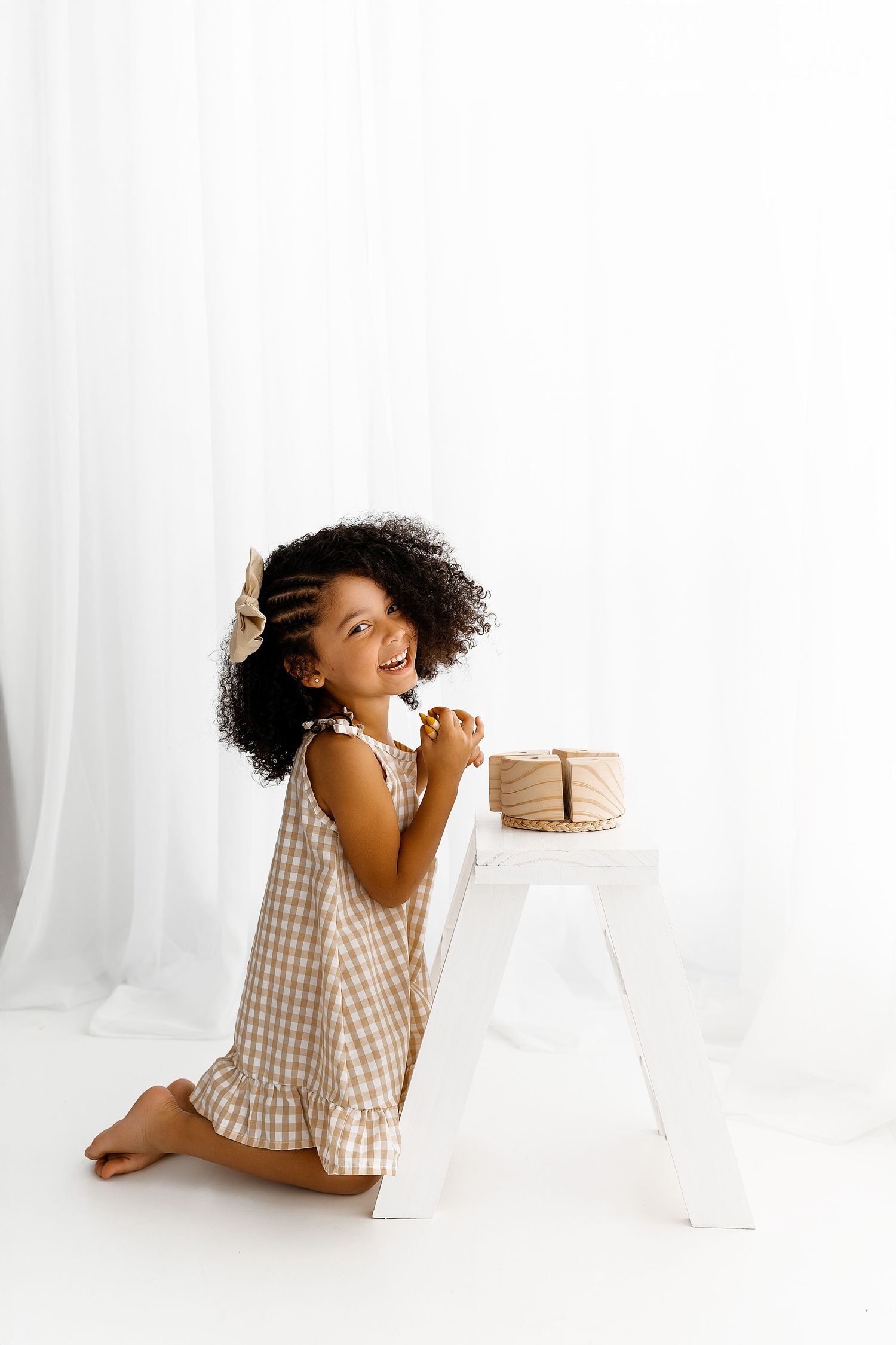 Young child in gingham dress kneeling beside white stool with small cake, against white backdrop.