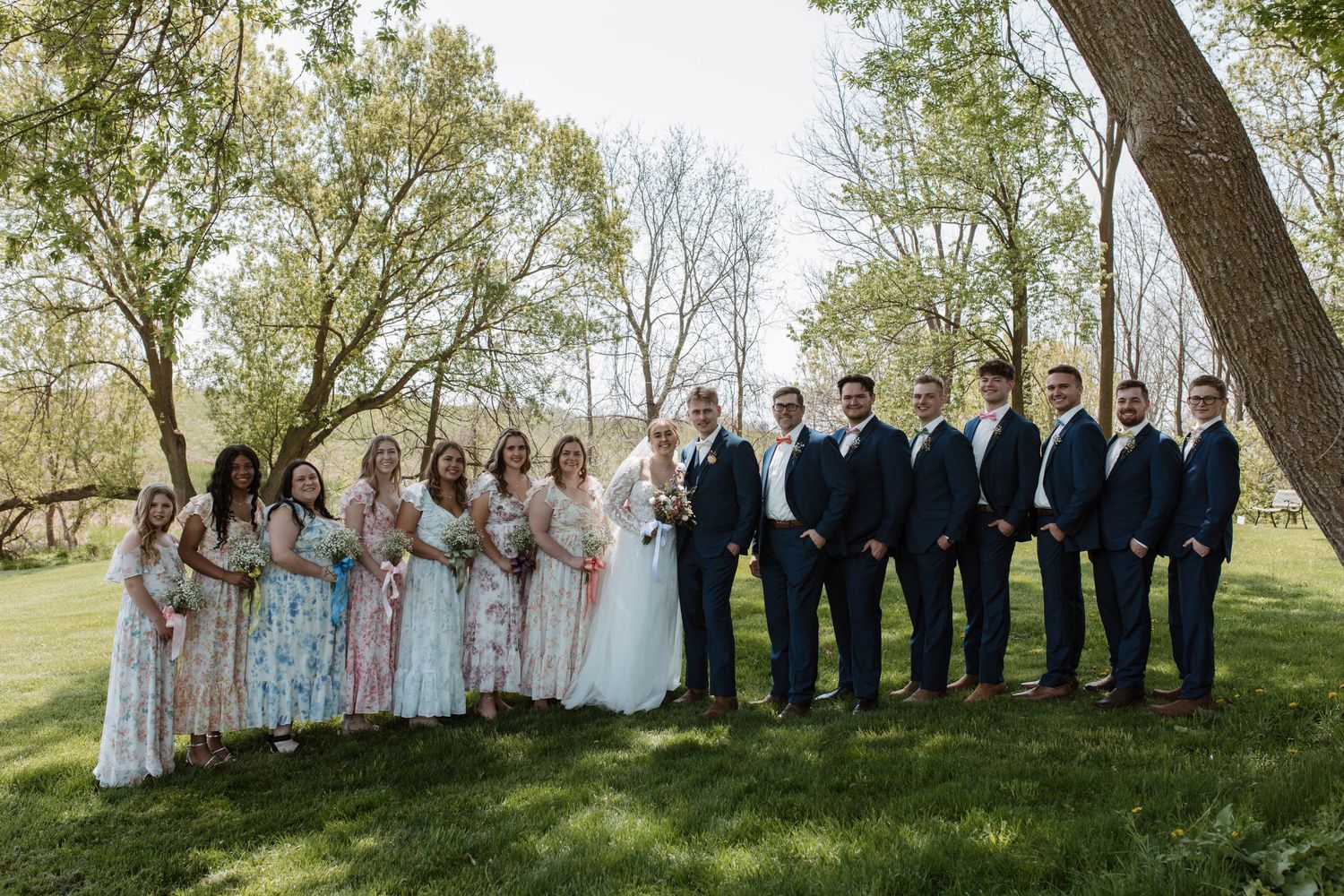 Large wedding party in floral bridesmaid dresses and navy groomsmen suits pose outdoors under spring trees.
