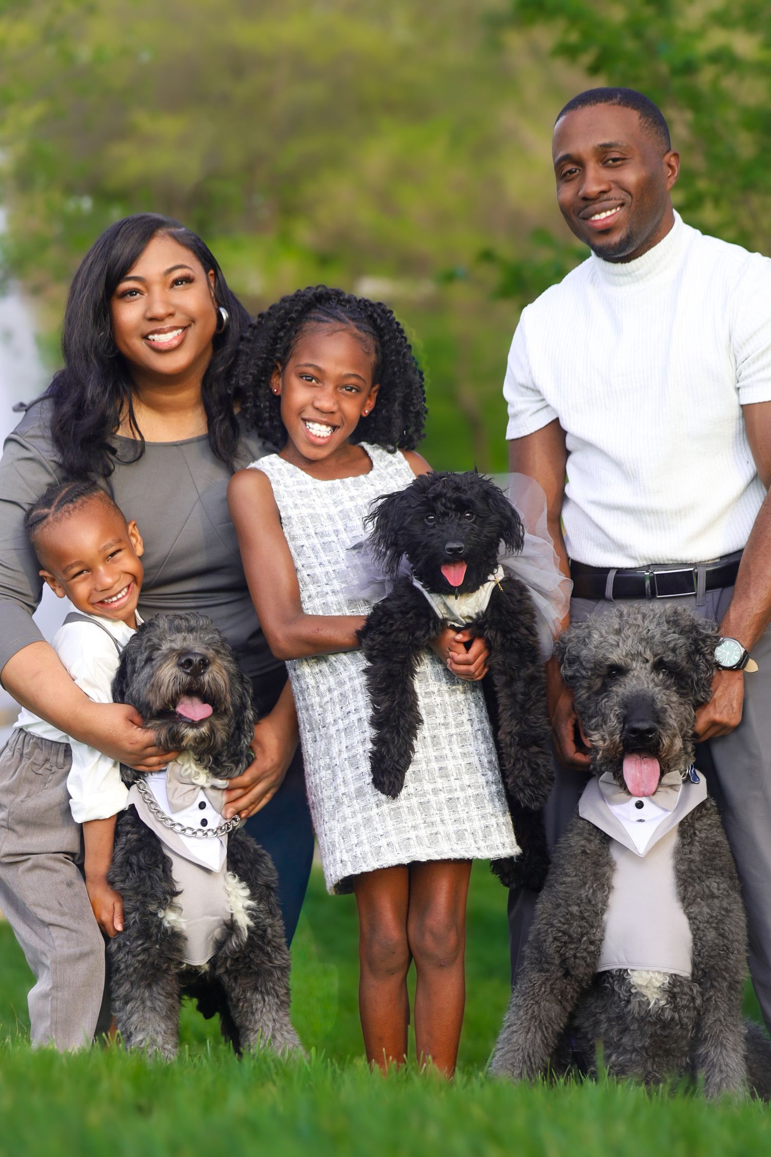 Happy Black family smiling outdoors on green grass with two fluffy black dogs in bowties.