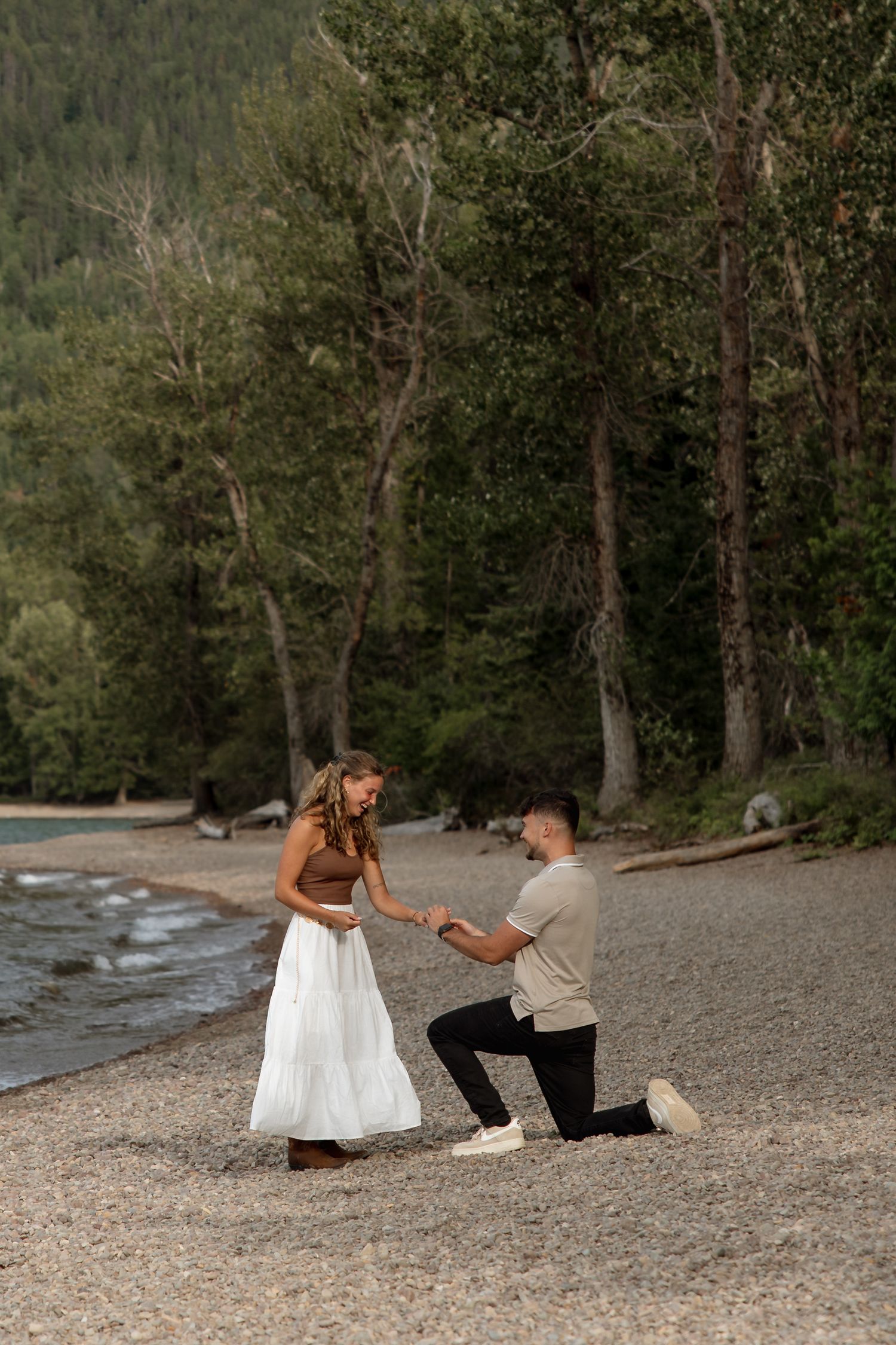 A couple embraces on a rocky lakeshore with mountains and autumn trees in the background.