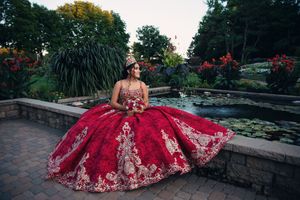 Ornate red ball gown with detailed embellishments displayed against a stone pathway and garden backdrop.