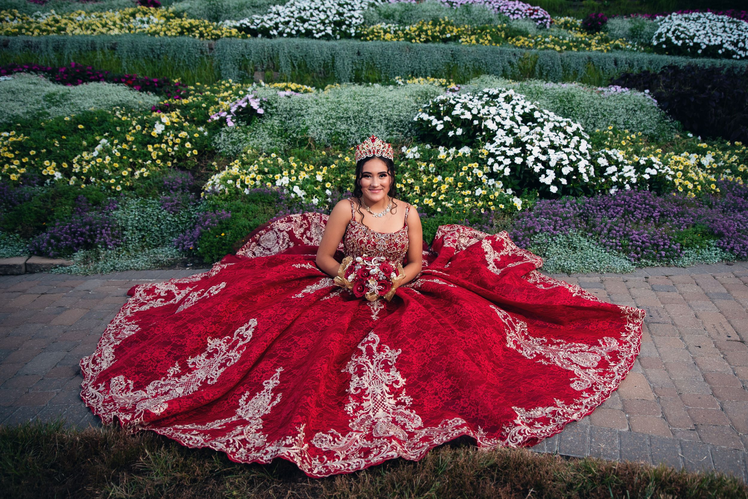 Luxurious crimson ball gown with metallic details photographed among colorful garden flowers.