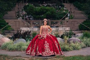 A vibrant red quinceañera gown with gold embellishments photographed in an outdoor garden setting.