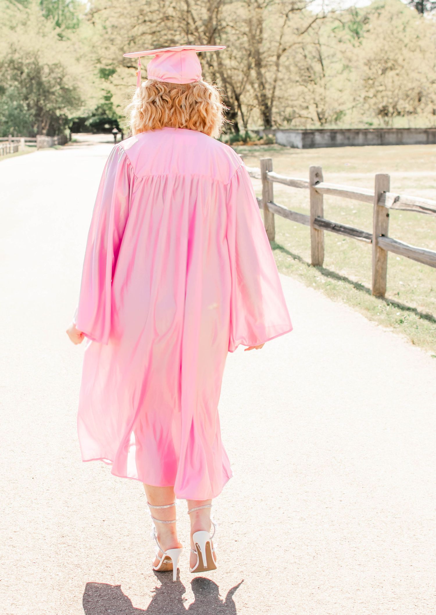Full length graduation photo showing flowing pink gown and wooden fence.