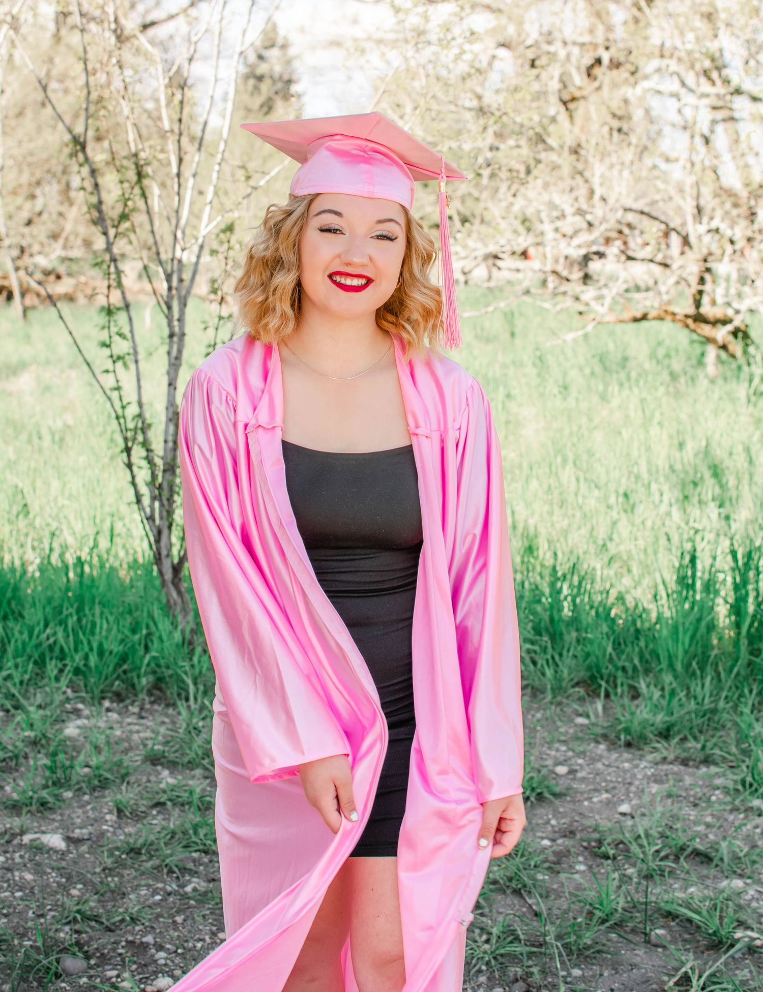 Graduate in pink cap and gown poses outdoors in natural spring setting.