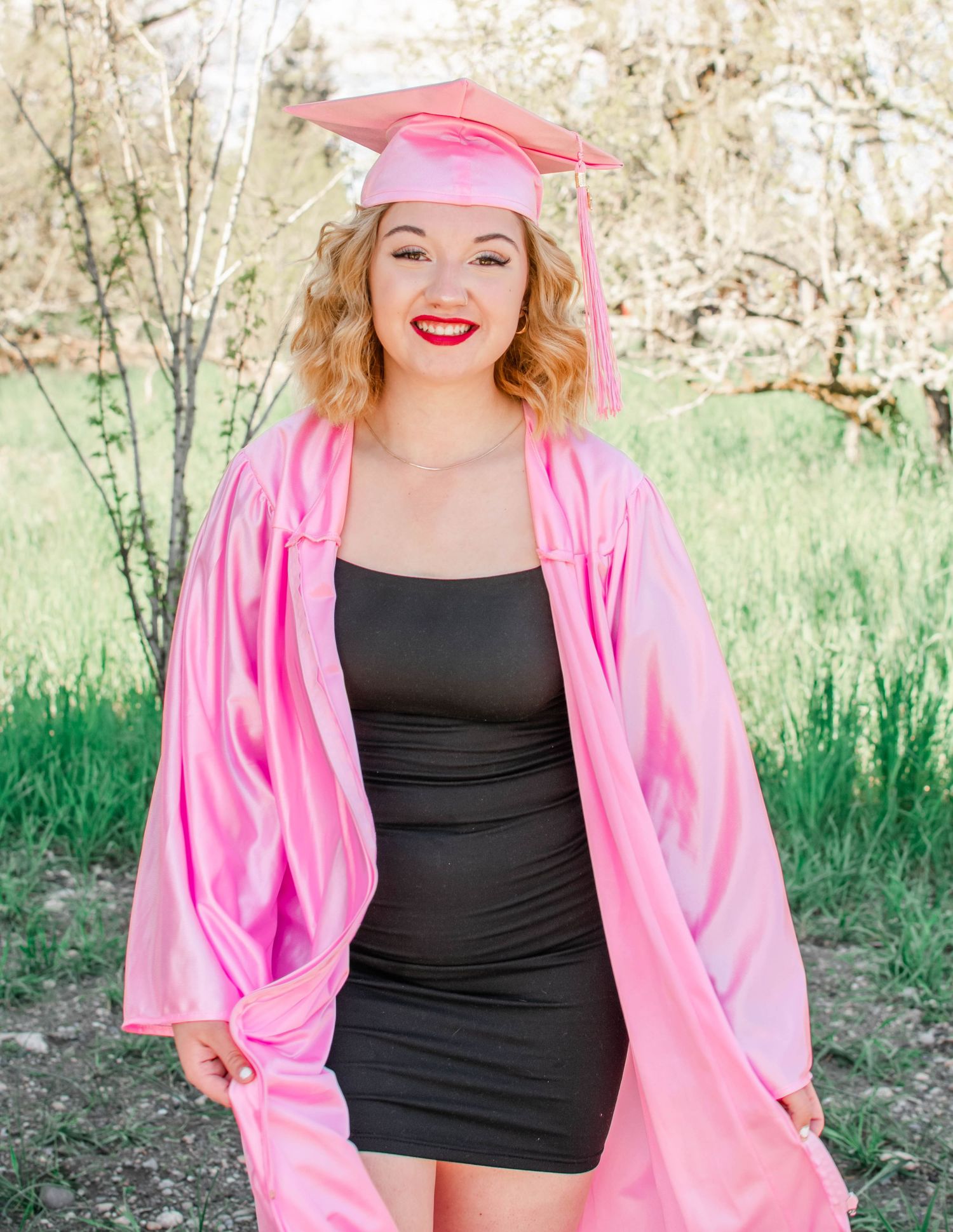 Graduate posing in a black dress and pink graduation gown against a natural outdoor background.