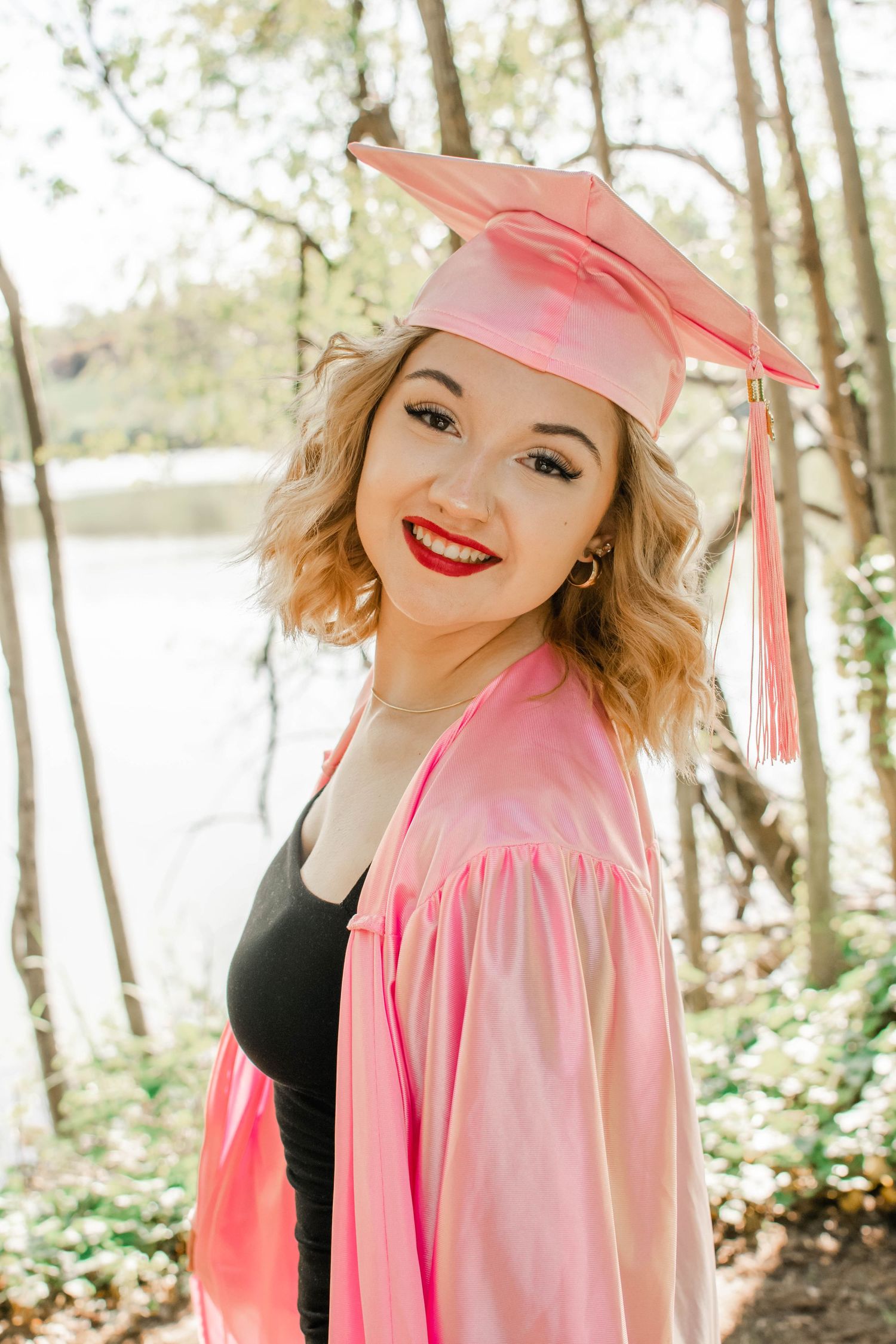 Pink graduation cap and gown portraits in a sunny wooded setting.