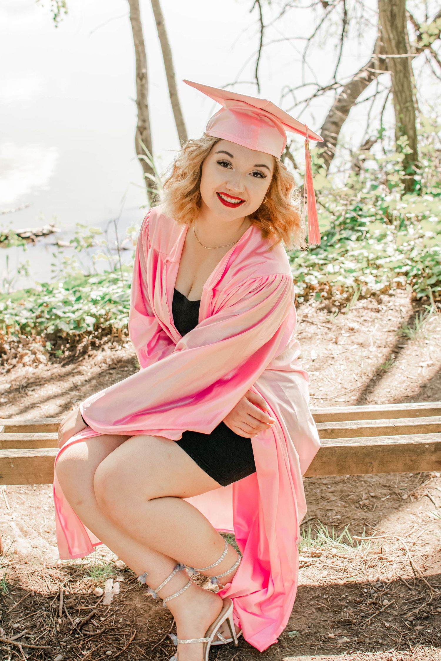 Graduation photoshoot featuring pink cap and gown on wooden bench in nature.
